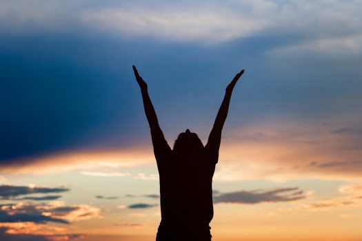 Happy celebrating winning success woman at sunset or sunrise standing elated with arms raised up above her head in celebration of having reached mountain top summit goal during hiking travel trek.