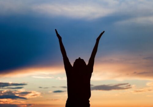 young woman silhouette at sunset Happy celebrating winning success woman at sunset or sunrise standing elated with arms raised up above her head in celebration of having reached mountain top summit goal during hiking travel trek.
