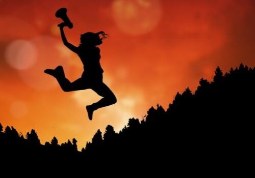 Businesswoman holding megaphone while jumping on mountain during sunset
